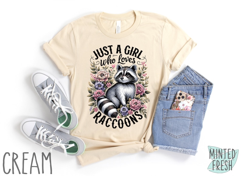 May include: A cream colored t-shirt with the text "Just a girl who loves raccoons" in black lettering. The text is surrounded by a floral design with a raccoon in the center.