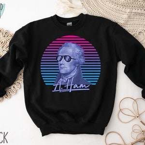 May include: Black sweatshirt featuring a retro-style graphic of Alexander Hamilton wearing sunglasses against a blue and pink striped background. The text "A. Ham" is below the graphic.