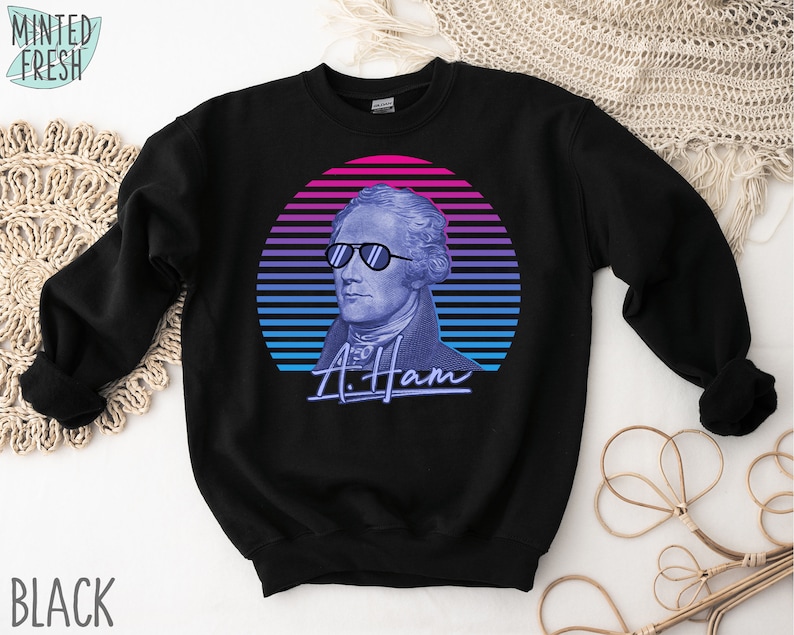 May include: Black sweatshirt featuring a retro-style graphic of Alexander Hamilton wearing sunglasses against a blue and pink striped background. The text "A. Ham" is below the graphic.