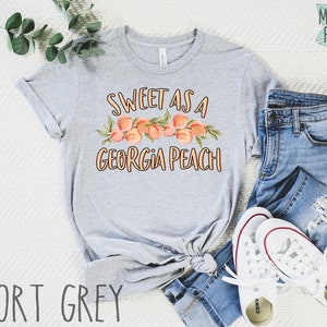 May include: A gray t-shirt with the text "Sweet as a Georgia Peach" and a graphic of peaches. The shirt is laid out on a white surface with a pair of blue jeans and white sneakers.
