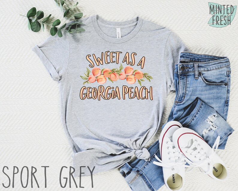 May include: A gray t-shirt with the text "Sweet as a Georgia Peach" and a graphic of peaches. The shirt is laid out on a white surface with a pair of blue jeans and white sneakers.