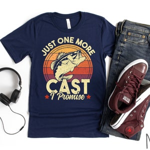 K&ouml;nnte beinhalten: Marineblaues T-Shirt mit einer Vintage-Grafik eines Fisches, der aus dem Wasser springt. Die Grafik ist von einem roten, orangefarbenen und gelben Sonnenuntergang umgeben. Der Text "Just One More Cast I Promise" ist unter der Grafik gedruckt.