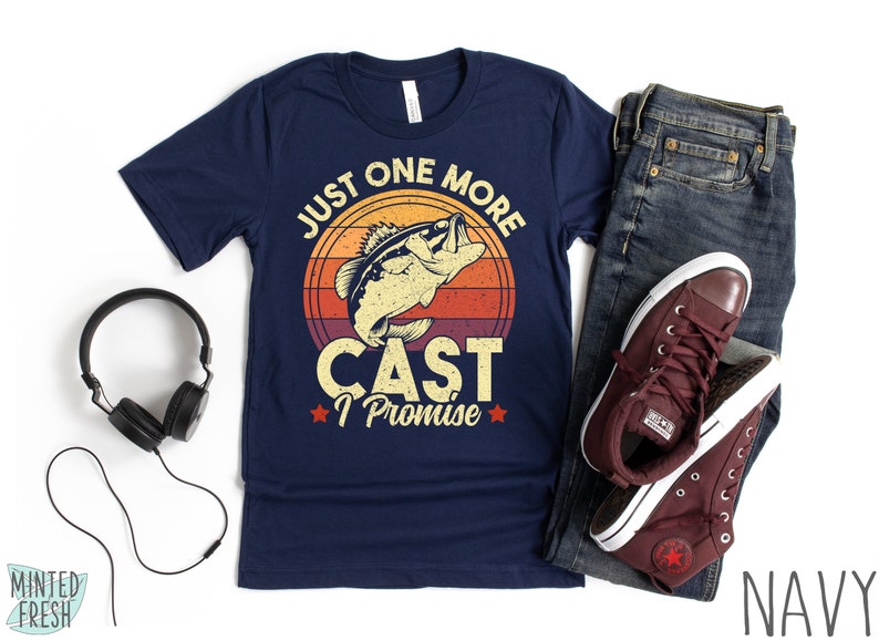 K&ouml;nnte beinhalten: Marineblaues T-Shirt mit einer Vintage-Grafik eines Fisches, der aus dem Wasser springt. Die Grafik ist von einem roten, orangefarbenen und gelben Sonnenuntergang umgeben. Der Text "Just One More Cast I Promise" ist unter der Grafik gedruckt.