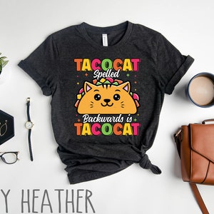 Puede incluir: Una camiseta gris jaspeada oscura con un gato de dibujos animados que lleva un taco. El texto de la camiseta dice "Tacocat Spelled Backwards is Tacocat".