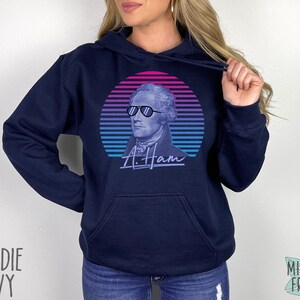 May include: A navy blue hoodie featuring a retro-style graphic of Alexander Hamilton wearing sunglasses. The graphic is in shades of pink, blue, and purple. The text "A. Ham" is printed below the graphic.