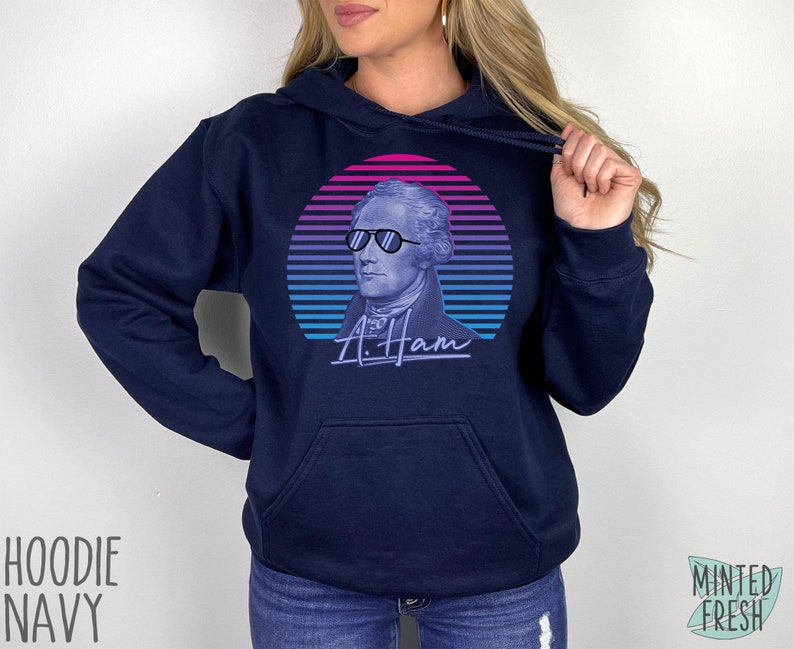 May include: A navy blue hoodie featuring a retro-style graphic of Alexander Hamilton wearing sunglasses. The graphic is in shades of pink, blue, and purple. The text "A. Ham" is printed below the graphic.