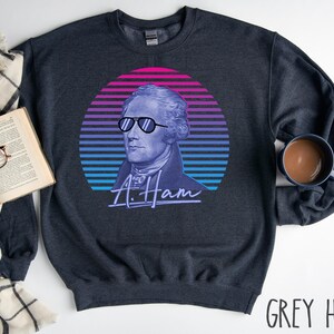 May include: A dark heather grey sweatshirt featuring a retro-style graphic of Alexander Hamilton wearing sunglasses. The graphic is set against a colourful sunset background with the text "A. Ham" underneath.