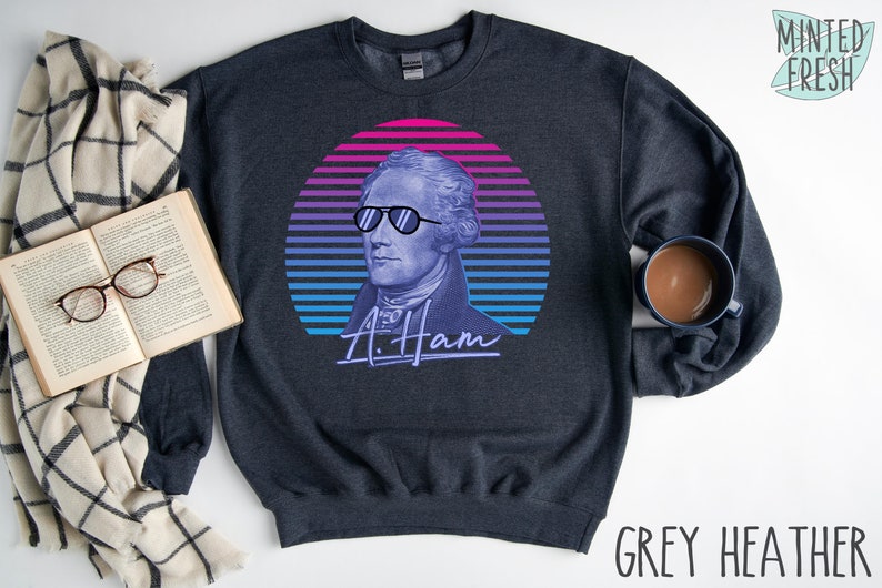May include: A dark heather grey sweatshirt featuring a retro-style graphic of Alexander Hamilton wearing sunglasses. The graphic is set against a colourful sunset background with the text "A. Ham" underneath.