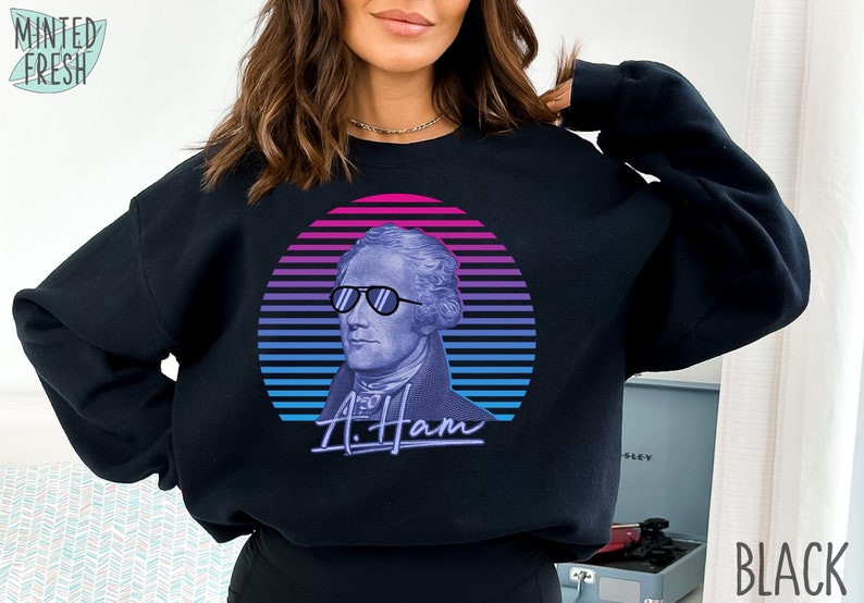 May include: Black sweatshirt featuring a retro-style graphic of Alexander Hamilton wearing sunglasses, with a blue and pink sunset behind him. The text "A. Ham" is written below the image.