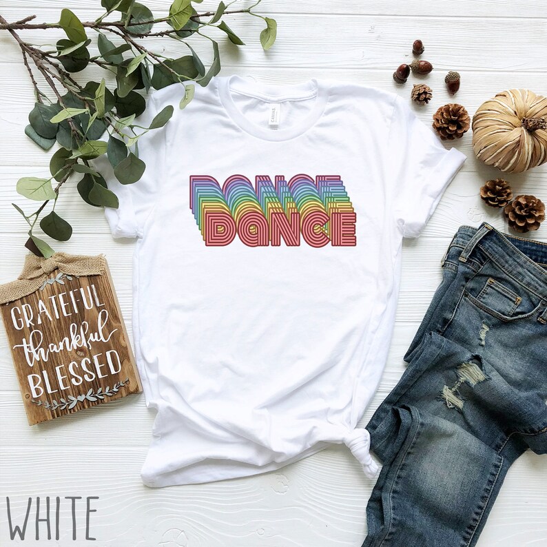 May include: White t-shirt with a rainbow-colored "DANCE" graphic. The shirt is laid flat on a white surface with a pair of blue jeans and a wooden sign that says "GRATEFUL thankful BLESSED".