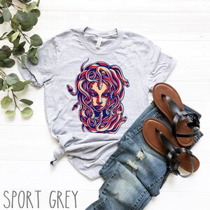 May include: A grey t-shirt with a graphic print of Medusa, a mythical Greek figure with snakes for hair. The snakes are red, blue, and yellow. The background of the graphic is white.