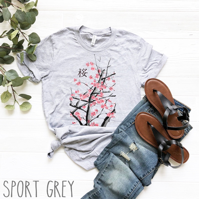 Puede incluir: Una camiseta gris con un dise&ntilde;o de &aacute;rbol de cerezo japon&eacute;s y la palabra "桜" impresa en la parte delantera. La camiseta est&aacute; colocada sobre una superficie blanca con un par de sandalias marrones y unos vaqueros azules.