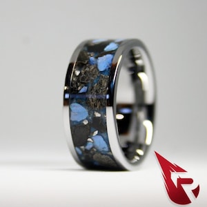 May include: A silver ring with a black and blue inlay. The inlay is made of small pieces of blue and black material that are set in a black resin. The ring is polished and has a smooth finish.