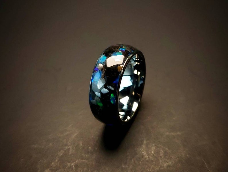 Puede incluir: Un anillo negro con una incrustaci&oacute;n azul y verde iridiscente. El anillo est&aacute; hecho de carburo de tungsteno y tiene un acabado pulido.