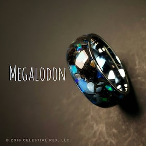 Puede incluir: Un anillo de carburo de tungsteno negro con una incrustaci&oacute;n moteada azul, verde y blanca. El anillo est&aacute; grabado con la palabra "MEGALODON".