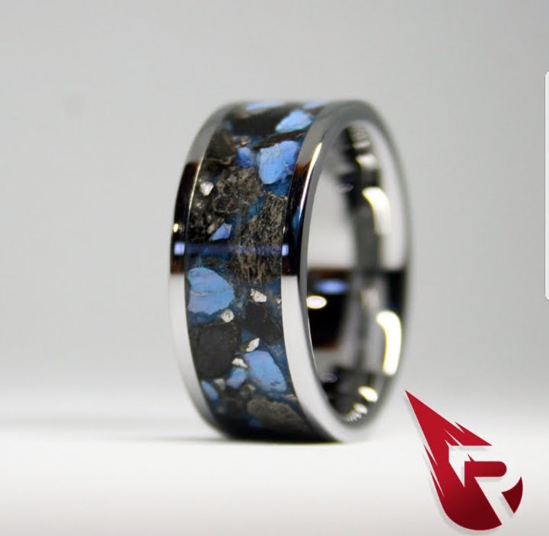 May include: A silver ring with a black and blue inlay. The inlay is made of small pieces of blue and black material that are set in a black resin. The ring is polished and has a smooth finish.