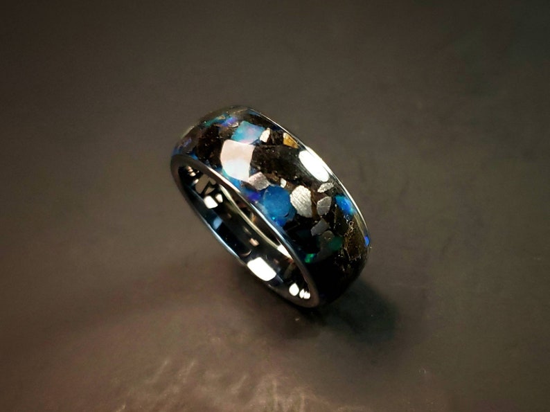 Puede incluir: Un anillo negro con una incrustaci&oacute;n azul y plateada. La incrustaci&oacute;n est&aacute; hecha de peque&ntilde;os trozos de material que est&aacute;n engastados en una resina.