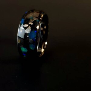 Puede incluir: Un anillo negro con una incrustaci&oacute;n azul y blanca. La incrustaci&oacute;n est&aacute; hecha de peque&ntilde;as piezas irregulares de material que est&aacute;n engastadas en el metal negro.