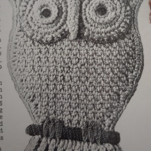 Op de afbeelding: Een gehaakt uilvormig item in grijstinten. De uil heeft grote, cirkelvormige ogen met donkere centra, een prominente snavel en een getextureerd lichaam. De onderkant heeft een geribbeld ontwerp en een donkere band.