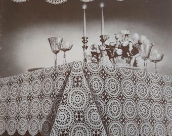 May include: A white crocheted tablecloth with a circular pattern. The tablecloth is laid out on a table with candlesticks and flowers in the center.