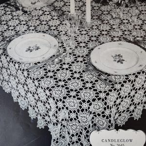 May include: A white crocheted tablecloth with a delicate pattern, set for a formal dinner with two place settings, candlesticks, and glasses. The tablecloth is labeled "CANDLEGLOW No. 7645 62 x 81 inches".