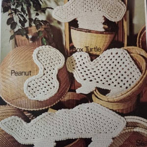 Puede incluir: Cuatro artículos de ganchillo en forma de hongo, cacahuete, tortuga de caja y tortuga marina. Los artículos son blancos y se muestran sobre cestas de mimbre.