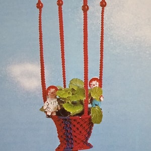 Könnte beinhalten: Ein roter und blauer Makramee-Pflanzenhänger mit einer weißen Kugel, die von oben hängt. Zwei kleine Puppen sitzen im Pflanzenhänger mit einer grünen Pflanze. Der Text "Up, Up And Away" ist unter dem Pflanzenhänger gedruckt.