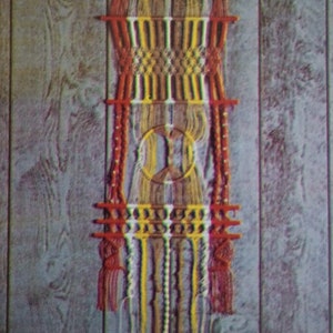 Op de afbeelding: Een kleurrijk macramé wandkleed met rood, geel, bruin en wit garen. Het kleed heeft een cirkelvormig ontwerp in het midden en een franje aan de onderkant.