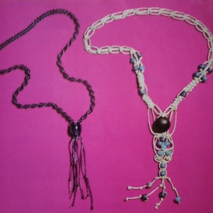 Op de afbeelding: Twee kettingen op een roze achtergrond. Eén ketting is zwart en heeft een kwast. De andere ketting is wit met blauwe kralen en een kwast.