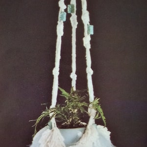 Puede incluir: Un macetero de macramé blanco con piedras verdes y un borla blanca. Una planta verde está en una maceta blanca colgando del macramé.