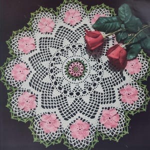 Könnte beinhalten: Eine weiße Häkeldeckchen mit grünem Rand und rosa Blumenakzenten. Die Deckchen liegt auf einer schwarzen Oberfläche mit zwei roten Rosen.