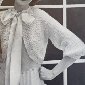 Puede incluir: Fotografía en blanco y negro de una mujer con un bolero blanco de punto con un lazo de satén. La mujer sonríe y posa cerca de una ventana. Imagen vintage.