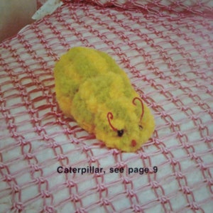Może przedstawiać: Żółto-zielona zabawka w kształcie gąsienicy na różowym kocyku robionym na szydełku. Koce ma wzór siatki, a pod zabawką nadrukowany jest tekst "Caterpillar, see page 9".