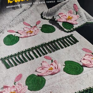 May include: A vintage crochet pattern booklet titled "Bathroom Beauties" featuring decorative crochet designs for bath towels, guest towels, and face cloths. The cover shows a white towel with pink and green crocheted water lily designs.