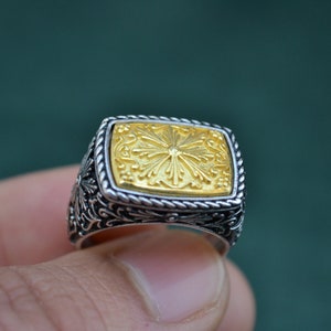May include: A silver ring with a gold square-shaped center piece featuring a floral design.