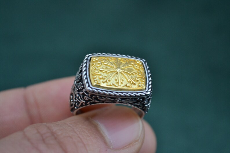 May include: A silver ring with a gold square-shaped center piece featuring a floral design.