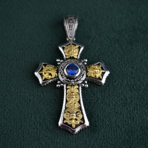 May include: A silver cross pendant with gold accents and a blue gemstone in the centre. The cross is intricately detailed with floral designs.