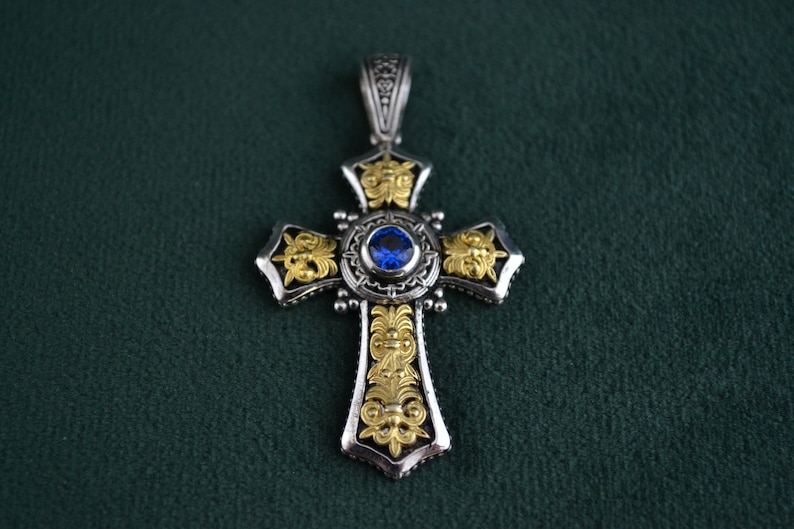 May include: A silver cross pendant with gold accents and a blue gemstone in the centre. The cross is intricately detailed with floral designs.