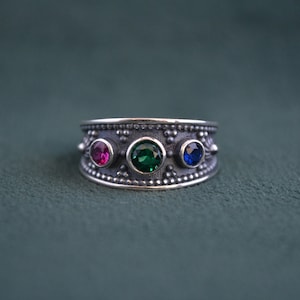 Op de afbeelding: Een zilveren ring met drie edelstenen: een roze steen, een groene steen en een blauwe steen. De ring heeft een getextureerde band.