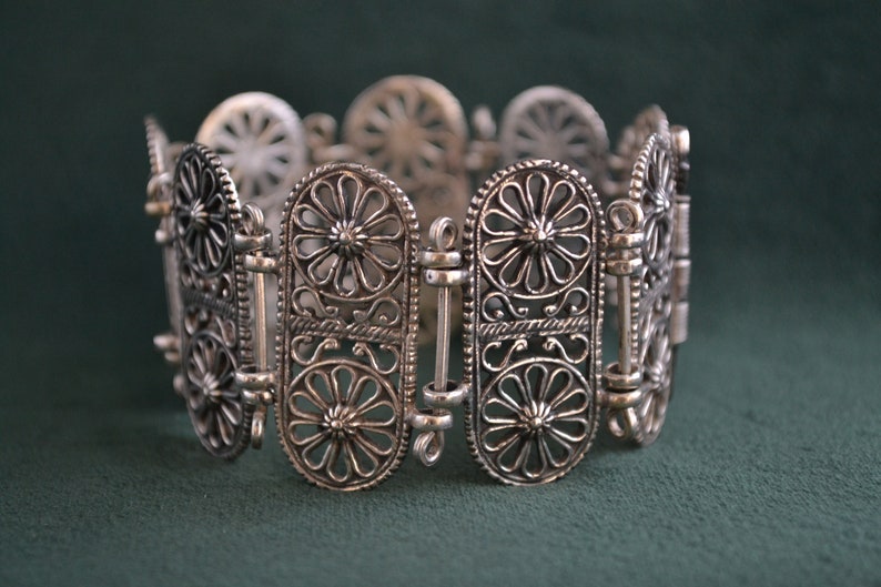 Puede incluir: Pulsera de filigrana plateada con un dise&ntilde;o calado. La pulsera presenta eslabones ovalados con motivos florales y bisagras de conexi&oacute;n. El intrincado trabajo en metal crea una apariencia delicada y ornamentada, adecuada para joyer&iacute;a.