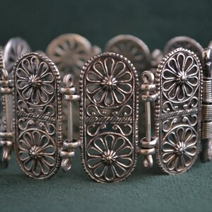 Puede incluir: Pulsera plateada con un intrincado dise&ntilde;o floral. La pulsera presenta eslabones ovalados con patrones de flores caladas y barras de conexi&oacute;n. El trabajo en metal es detallado y la pulsera est&aacute; sobre un fondo verde oscuro.