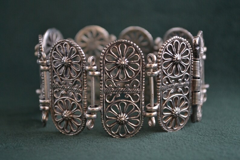Puede incluir: Pulsera plateada con un intrincado dise&ntilde;o floral. La pulsera presenta eslabones ovalados con patrones de flores caladas y barras de conexi&oacute;n. El trabajo en metal es detallado y la pulsera est&aacute; sobre un fondo verde oscuro.