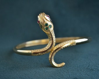 May include: A gold snake bracelet with green and red gemstones. The snake is coiled and the head is raised. The bracelet is on a dark blue background.