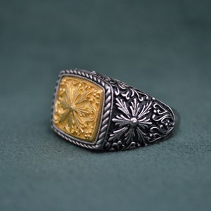 May include: A silver ring with a gold square-shaped center featuring an intricate floral design. The ring has a detailed floral pattern around the band.