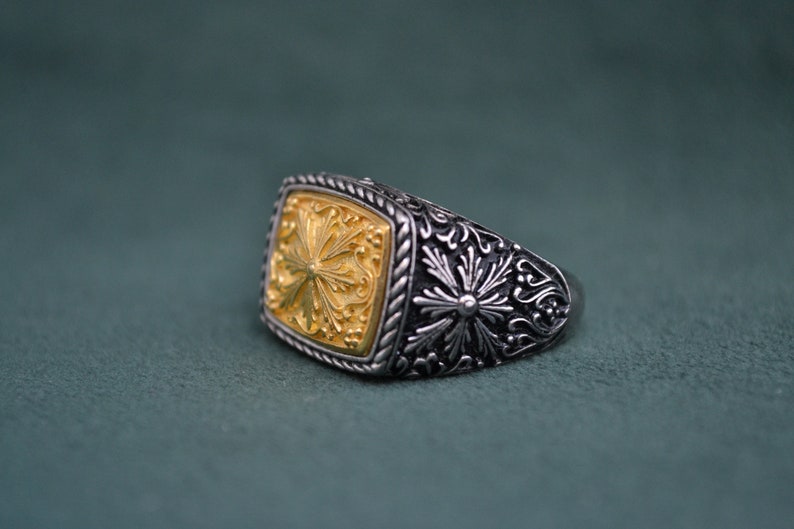 May include: A silver ring with a gold square-shaped center featuring an intricate floral design. The ring has a detailed floral pattern around the band.