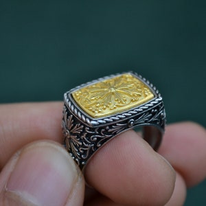 May include: A silver ring with a gold square-shaped top featuring an intricate floral design.
