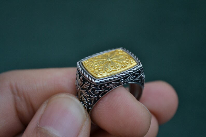May include: A silver ring with a gold square-shaped top featuring an intricate floral design.