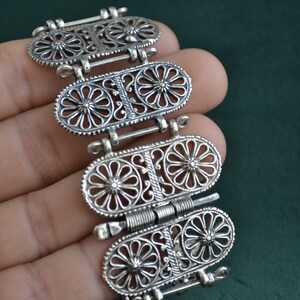 Puede incluir: Un brazalete plateado con un dise&ntilde;o ornamentado. El brazalete presenta cuatro eslabones ovalados, cada uno adornado con un patr&oacute;n floral y conectados por bisagras. Los intrincados detalles y el trabajo de filigrana le dan una apariencia vintage o antigua.