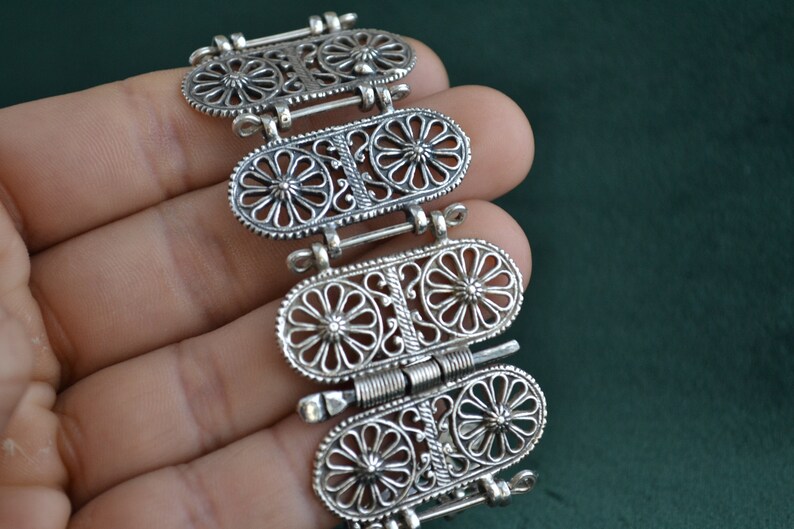 Puede incluir: Un brazalete plateado con un dise&ntilde;o ornamentado. El brazalete presenta cuatro eslabones ovalados, cada uno adornado con un patr&oacute;n floral y conectados por bisagras. Los intrincados detalles y el trabajo de filigrana le dan una apariencia vintage o antigua.