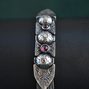 May include: A silver bracelet with a chain design and a series of oval-shaped links. Each link has a raised floral pattern and a red gemstone in the center.